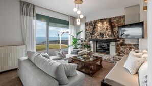 A modern living room with a stone fireplace, a large TV, and a view of the sea. - Villa Eagles - With Private Pool (Rethymno)
