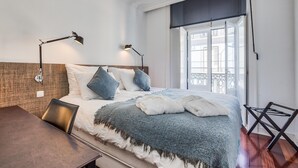 A bedroom with a bed, two bedside lamps, a window with a view, and a wooden floor. - Nomad s Chiado Lisbon - The Ivens Signature (Lisbon)