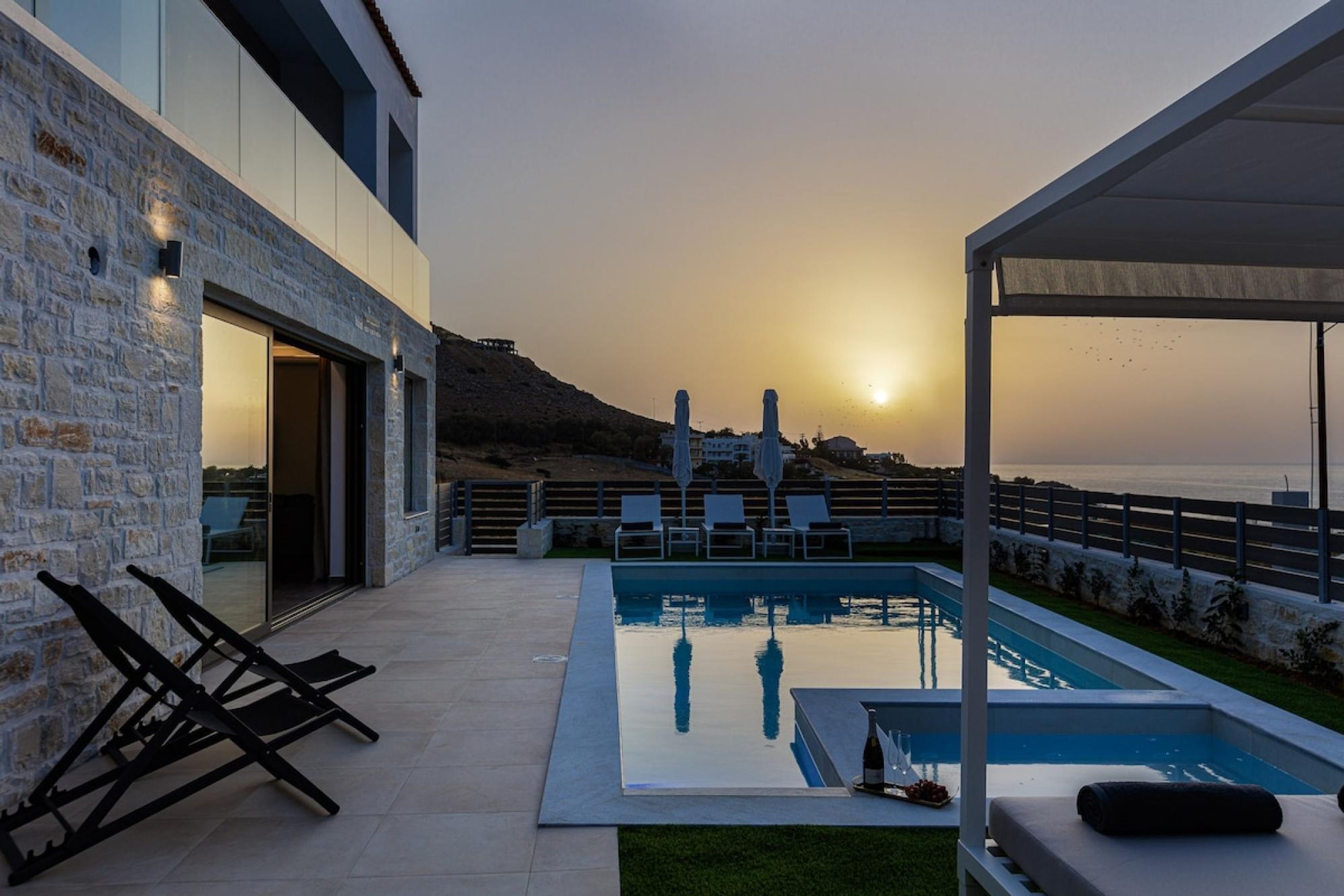 Ein moderner Poolbereich im Freien mit einer Steinmauer, Liegestühlen und Blick auf das Meer bei Sonnenuntergang.