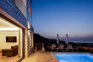 A modern outdoor pool area with lounge chairs and a TV screen, overlooking a scenic coastal view. - Dodici Luxury Villa - With Heated Pool (Mylopotamos)