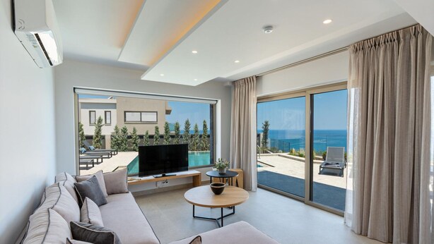 A modern living room with a sofa, a coffee table, a flat-screen TV, and a large window overlooking a pool and the sea. - Luxury Villa Aqua With 2 Private Pools (Rethymno)