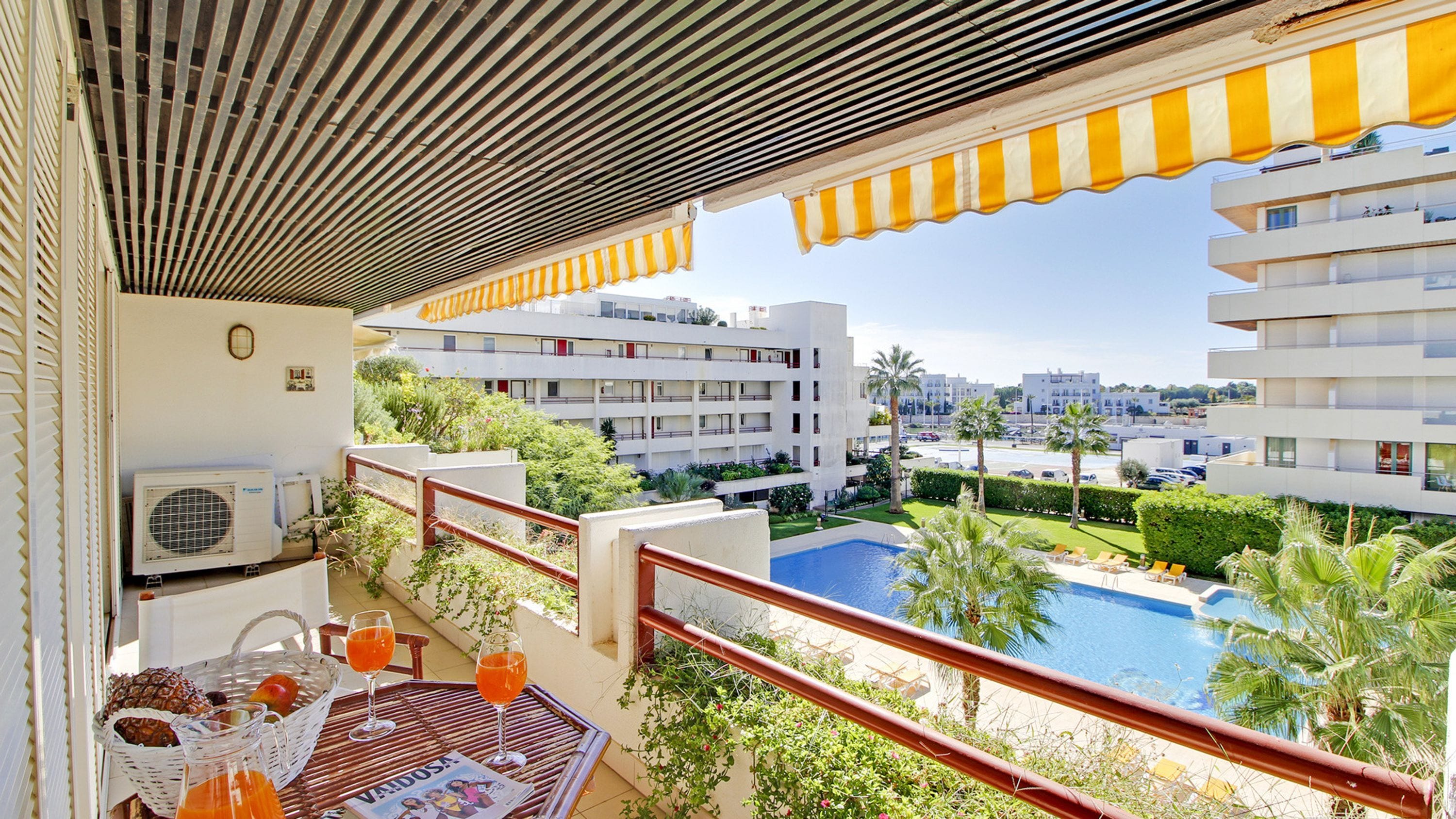 Varanda com vista para uma piscina, mesa com bebidas e um jornal, e um edifício ao fundo.