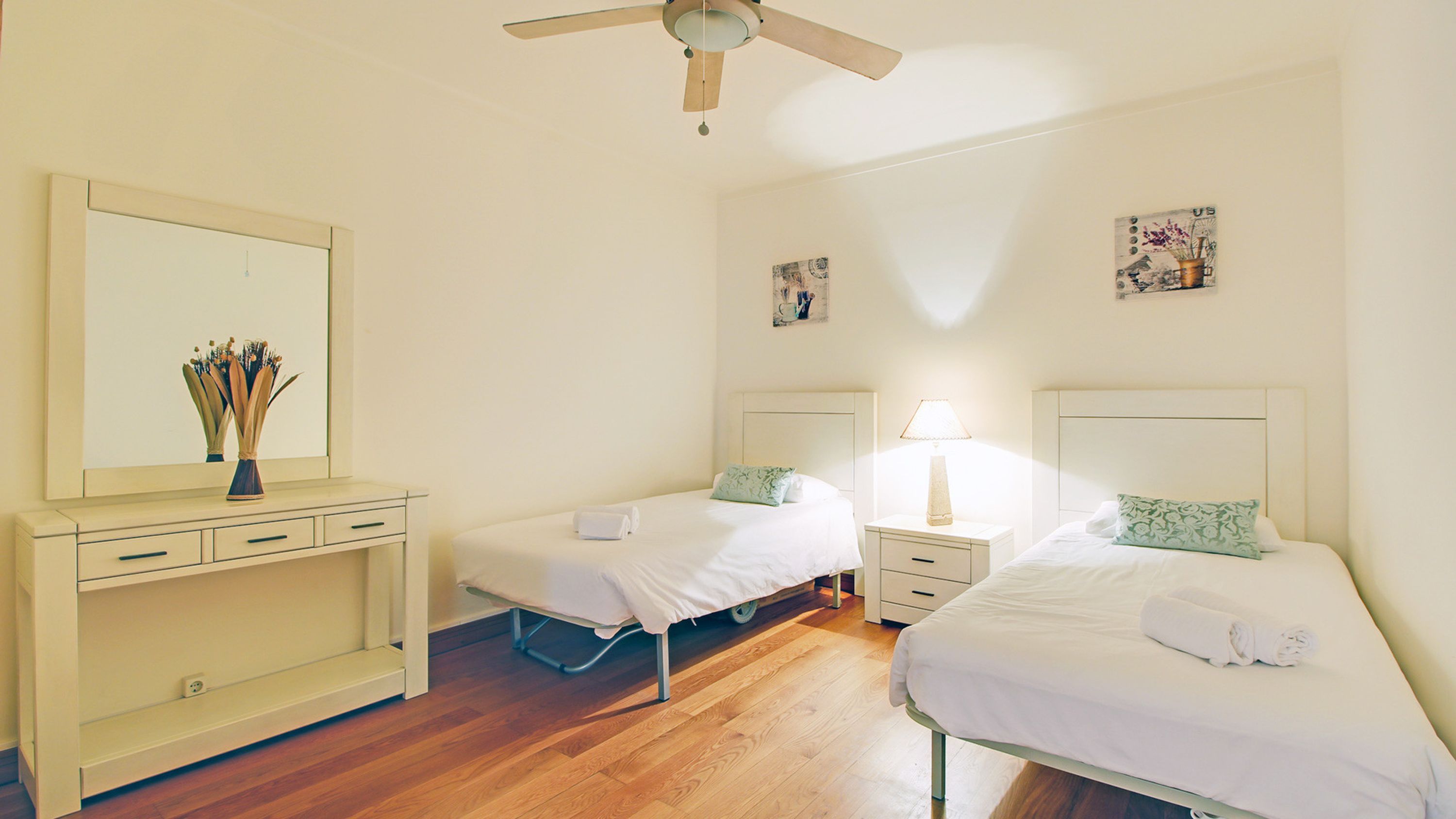 A hotel room with two single beds, a bedside table with a lamp, a dresser, and a ceiling fan.