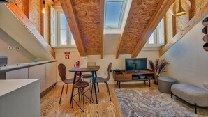 Une cuisine moderne avec des poutres en bois, un coin repas avec des chaises et une table, un téléviseur, et un petit coin salon avec un tapis et des plantes décoratives.