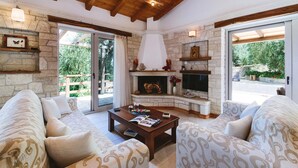 A cozy living room with stone walls, a fireplace, and a large window overlooking greenery. - Aristea - 2 BR Villa Surrounded by Olive Groves (Paxos)