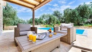 An outdoor seating area with wicker furniture, a table with breakfast items, and a view of a swimming pool. - Aristea - 2 BR Villa Surrounded by Olive Groves (Paxos)