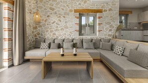 A modern living room with a stone wall, a wooden coffee table, and a grey sectional sofa with patterned cushions. - Armonia II Villa in Paxi (Paxos)