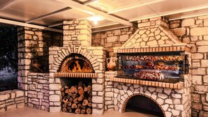 Cocina exterior de piedra con horno de leña, una parrilla y una vitrina con comida.