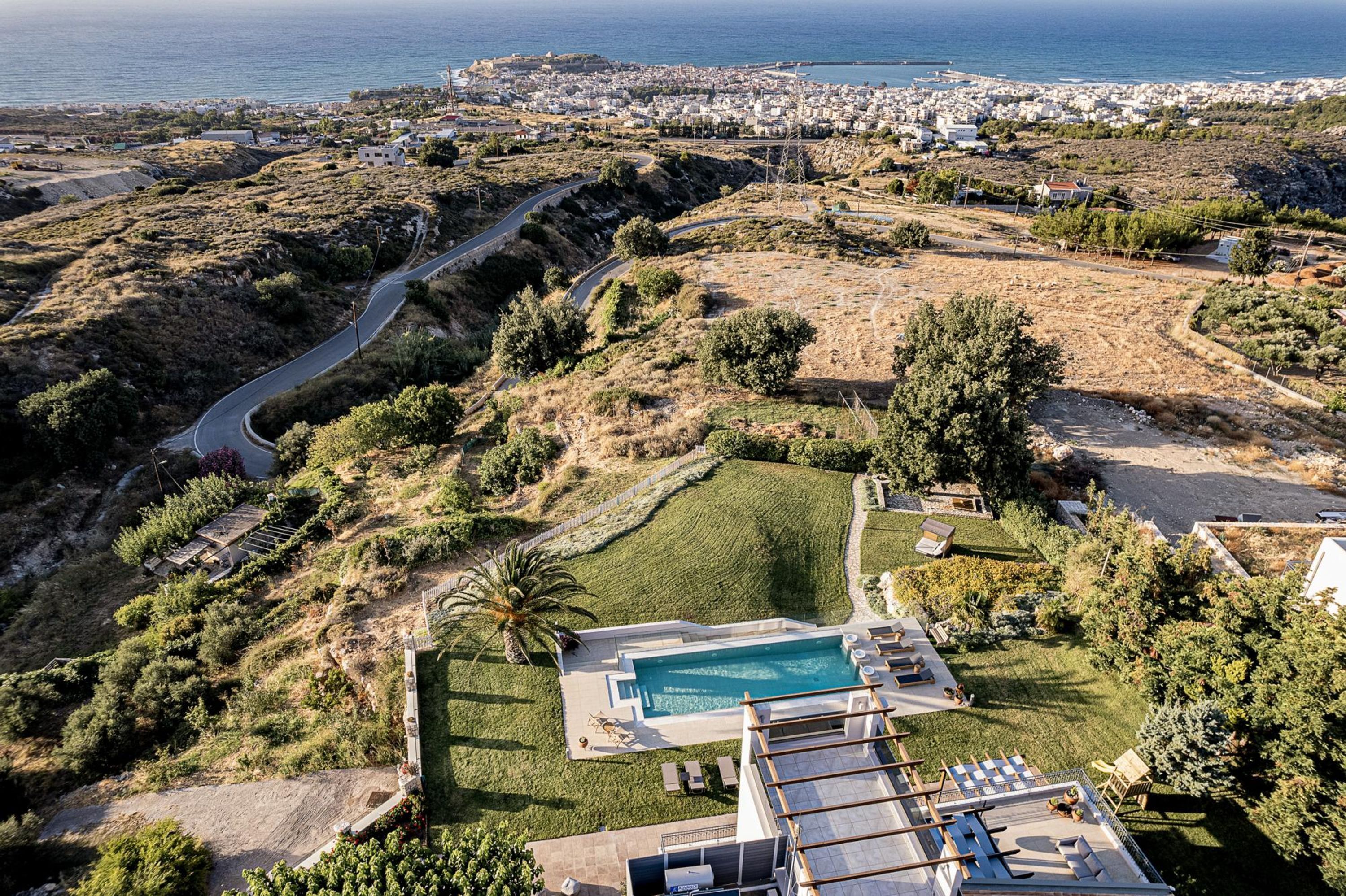 An aerial view of a property with a swimming pool, a winding road, and a view of the sea and town in the background.
