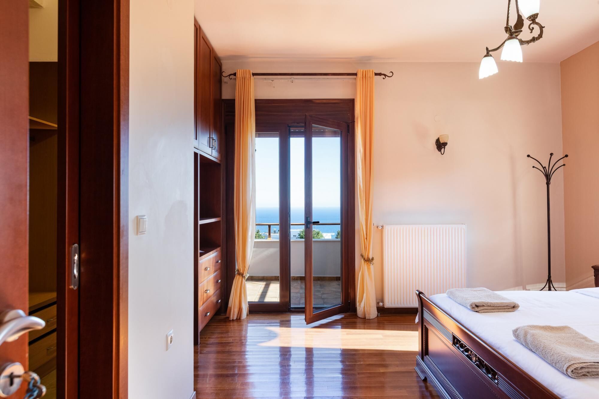 A bedroom with a bed, a radiator, a wardrobe, and a view of the sea through a glass door.