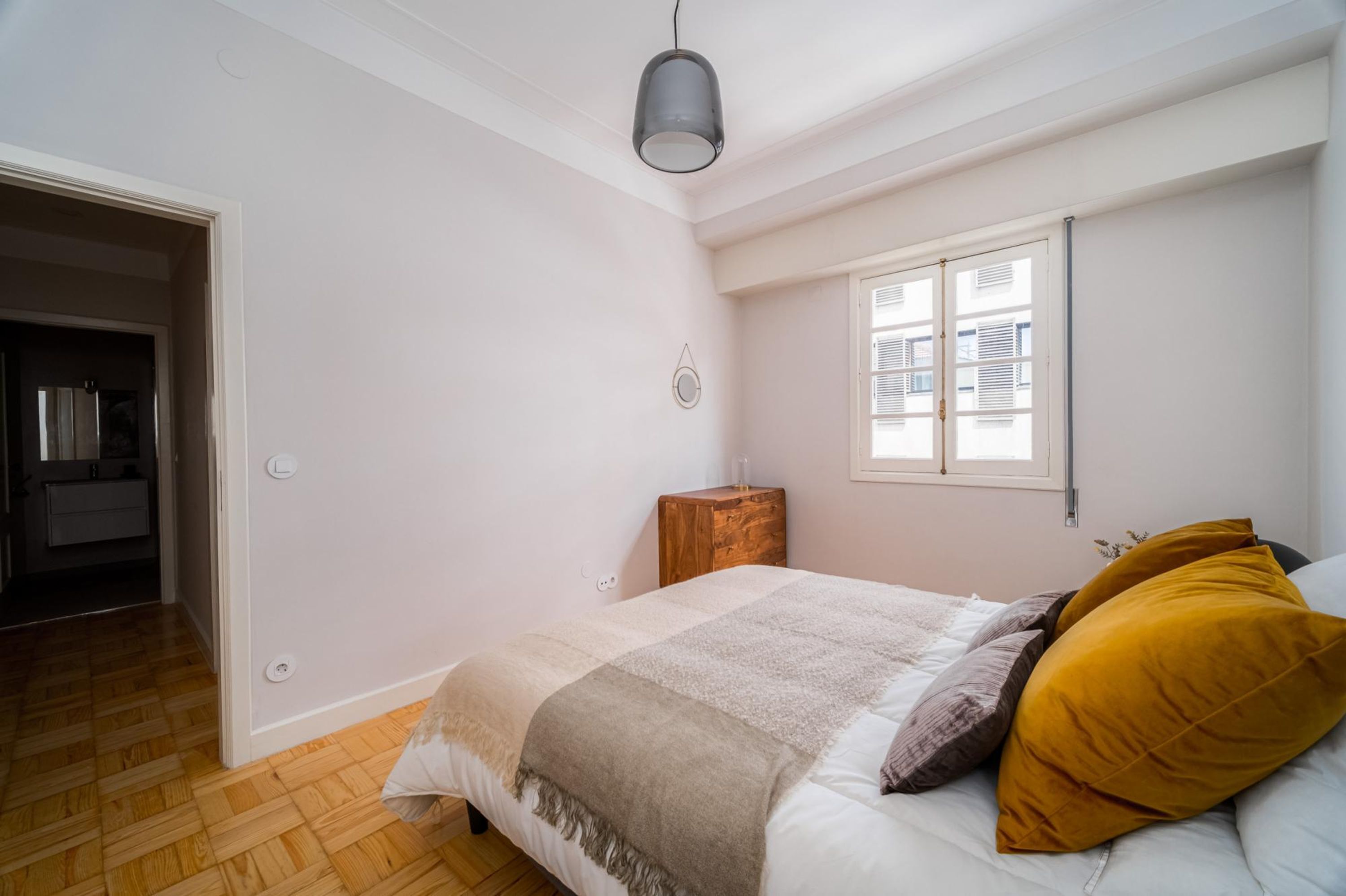 A bedroom with a bed, a bedside table, a window, and a door leading to another room.
