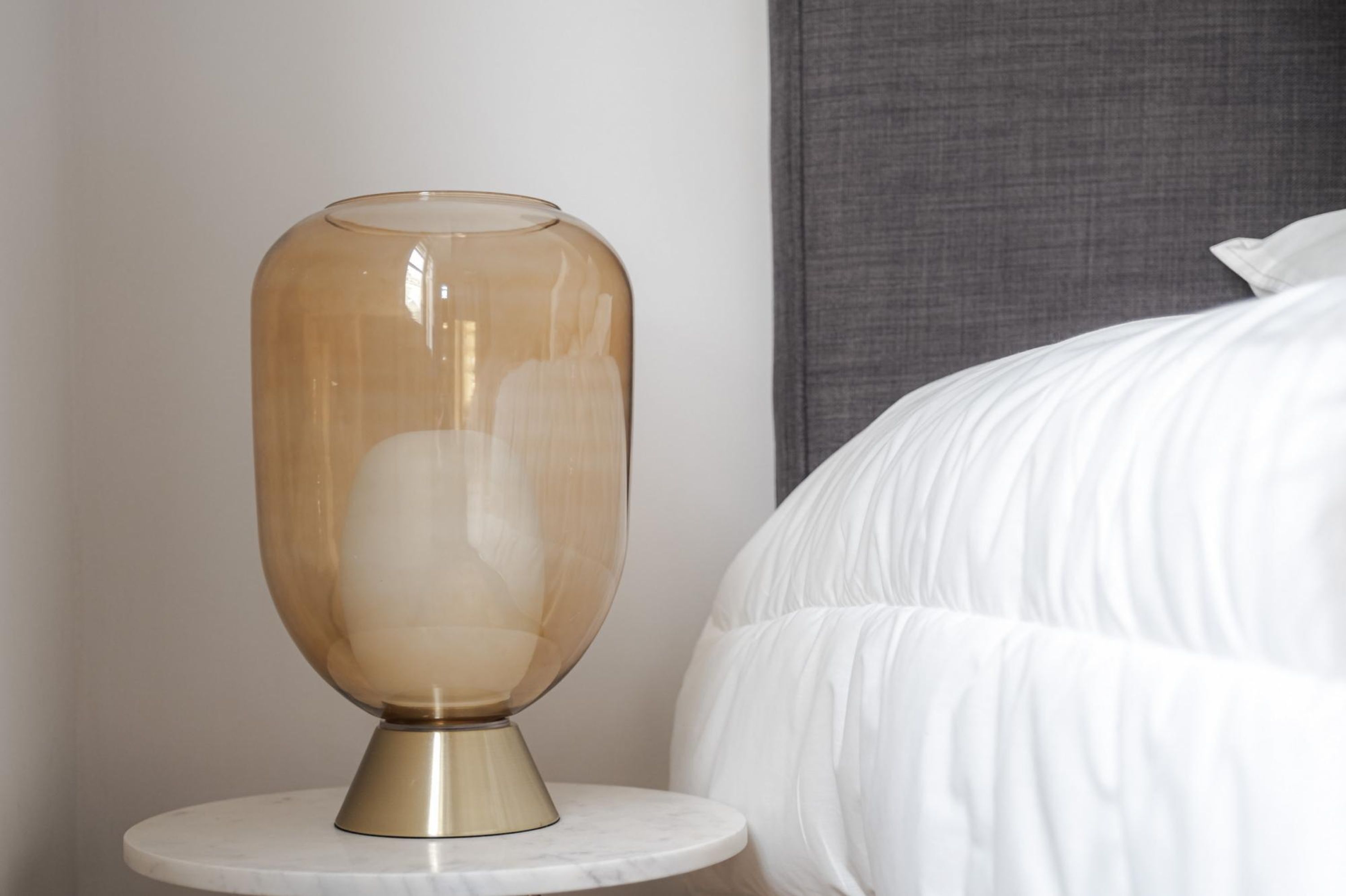 A bedside table with a clear glass lamp and a white bed.