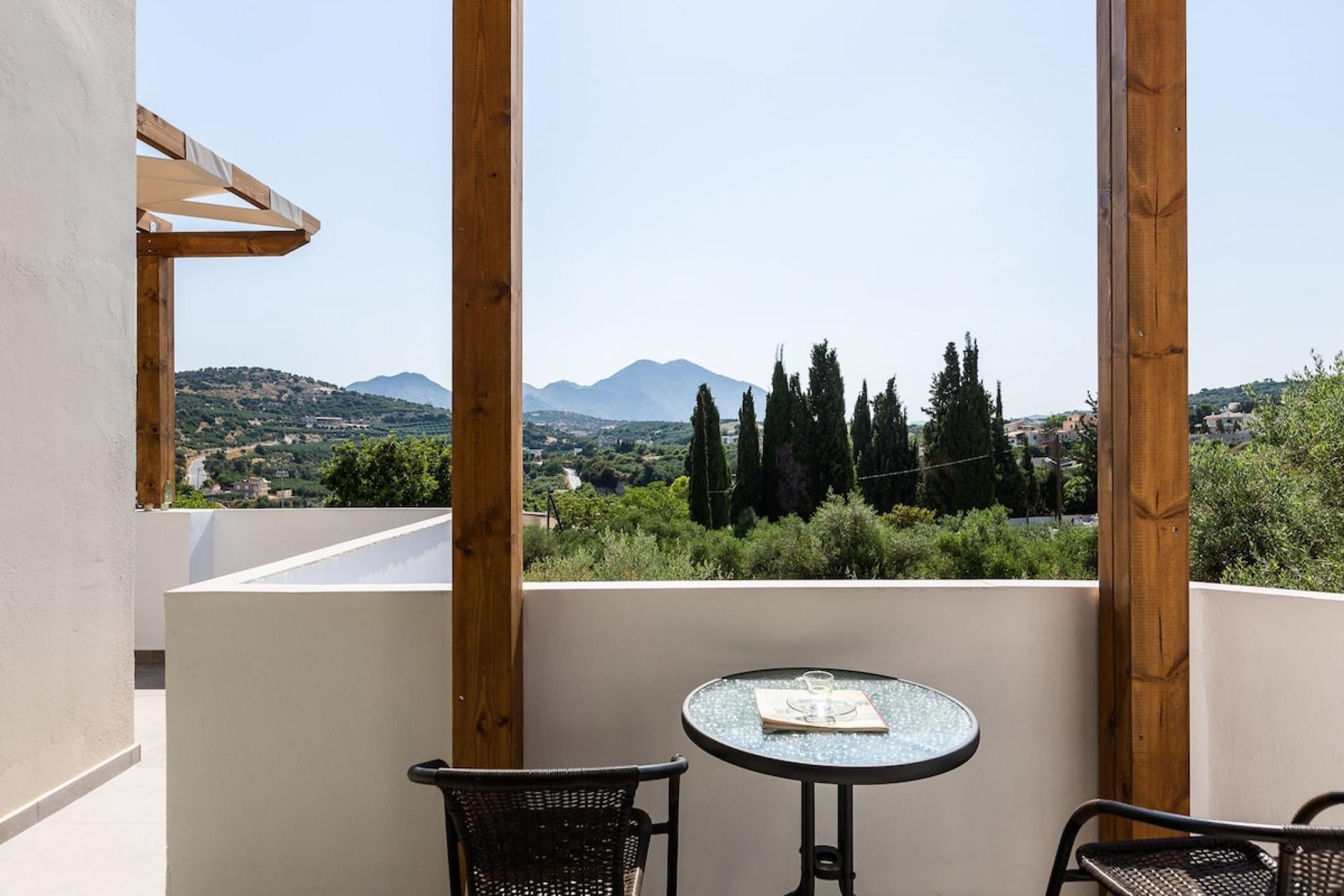 Un balcon avec une table et des chaises, donnant sur un paysage montagneux parsemé d’arbres et de bâtiments.