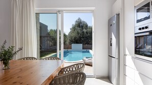 Een moderne keuken met een inbouwoven en koelkast, een eethoek met een houten tafel en rieten stoelen, en een schuifdeur die leidt naar een achtertuin met een zwembad.