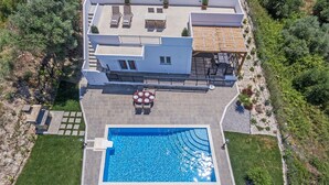 An aerial view of a modern house with a swimming pool, patio with outdoor furniture, and a fenced garden area. - Villa Guinevere-with Private Pool (Rethymno)