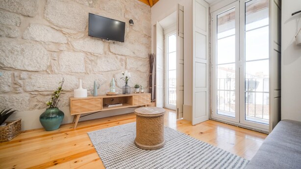 A room with a stone wall, wooden floor, a flat-screen TV, a wooden cabinet, a grey sofa, and a large window with a view. - Bonjardim City Flats - Top Floor (Porto)
