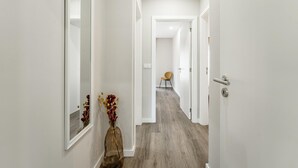 Een smalle gang met witte muren en een houten vloer, leidend naar een kamer met een stoel en een deur.