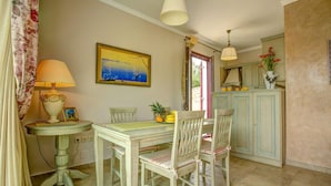 A dining area with a table and chairs, a lamp, a painting, and a kitchen area with cabinets and a vase of flowers. - Kalliope Villa - Classic Modern With a Splash of Glam (Paxos)