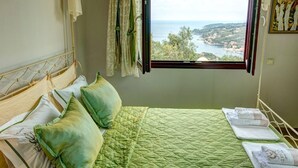 Een slaapkamer met een bed, kussens, een sprei, een raam met uitzicht op water en groen, en een schilderij aan de muur.