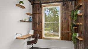 Una habitación con un ventanal grande, estantes de madera con plantas, un escritorio y una silla, y una pared blanca.