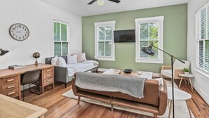 Ein modernes Schlafzimmer mit Bett, Schreibtisch und Sitzgelegenheit, ausgestattet mit Deckenventilator, großen Fenstern und einem Fernseher.