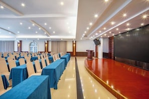 A conference room with rows of tables and chairs, a stage with a podium, and a large screen.
