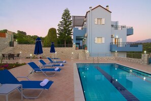 A pool area with lounge chairs and umbrellas, a stone wall, and a two-story building with a balcony. - Blue Villa 2 - With Private Pool (Mylopotamos)
