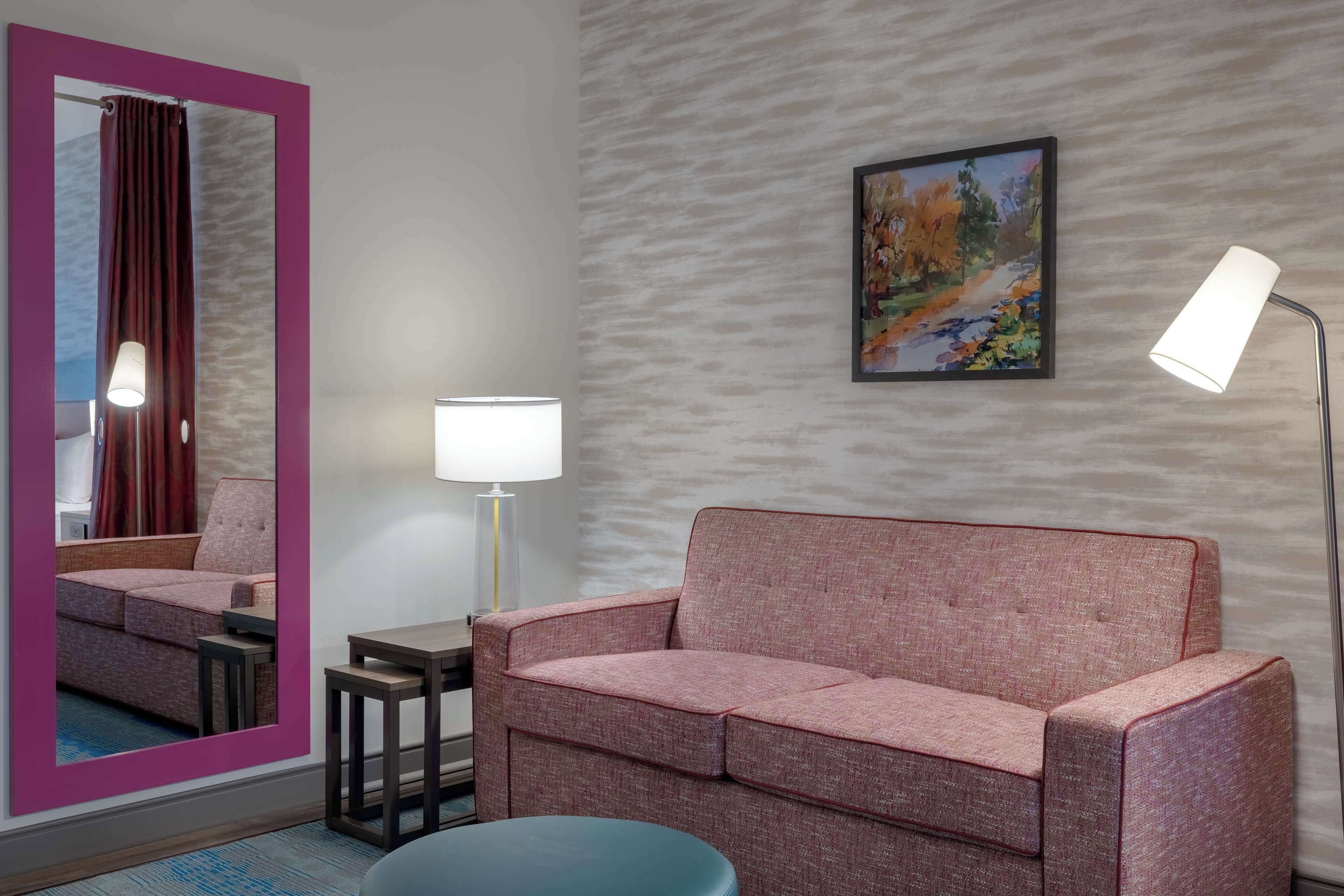 A modern hotel room with a pink sofa, a small table, a floor lamp, a mirror, and a painting on the wall.