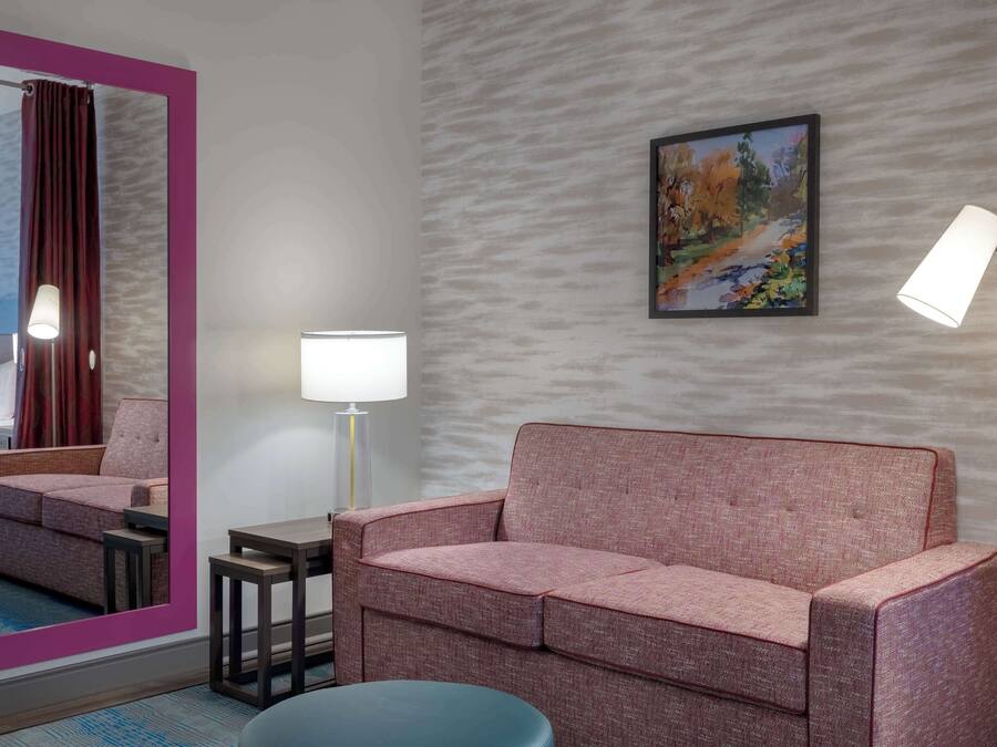 Una habitación de hotel moderna con un sofá rosa, una mesita, una lámpara de pie, un espejo y un cuadro en la pared.