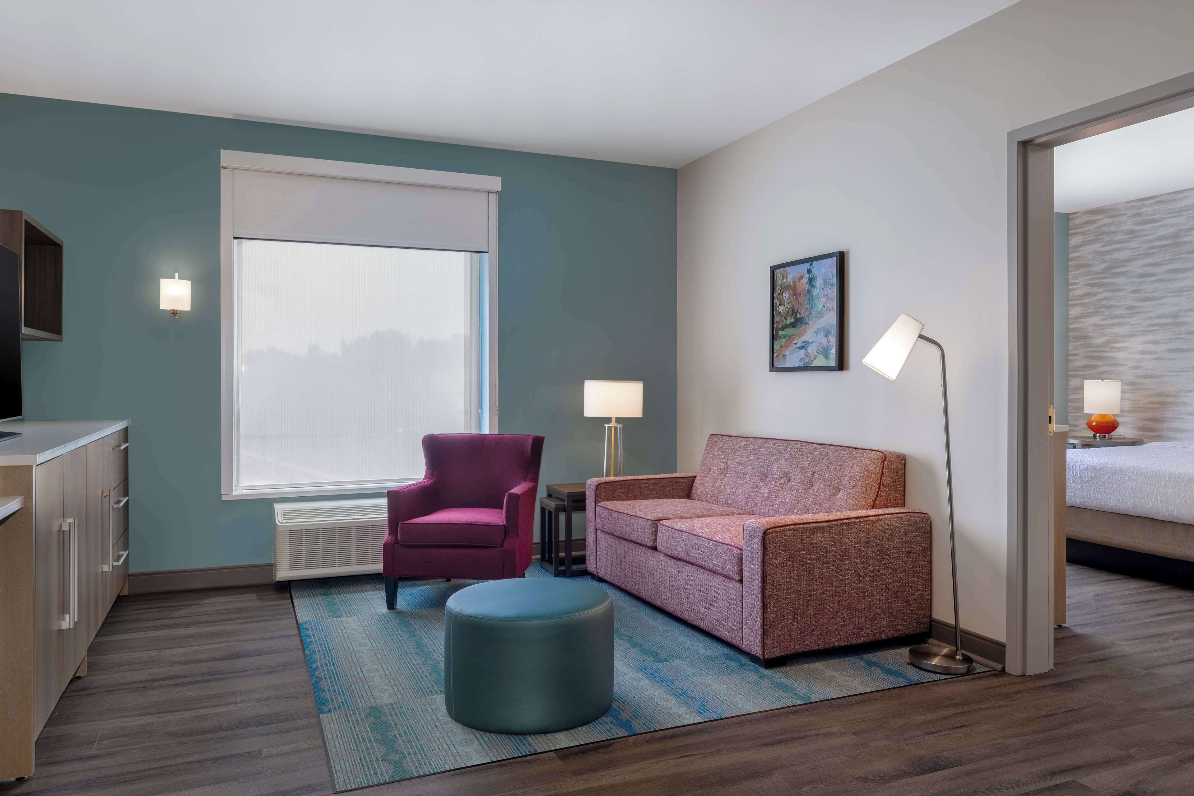 A hotel room with a kitchenette, a purple armchair, a pink sofa, a round ottoman, a large window with blinds, a painting on the wall, and a bed with a white headboard.