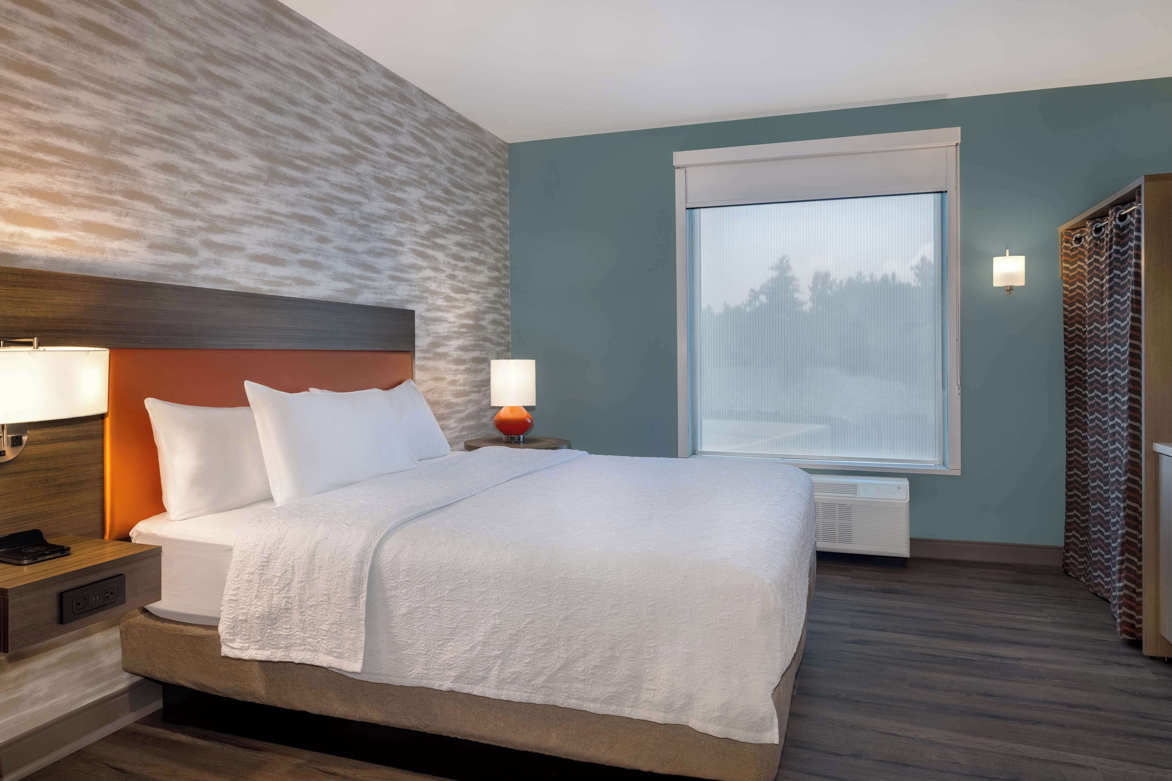 A hotel room with a bed, nightstands, a wall-mounted lamp, a window with blinds, and a wardrobe.