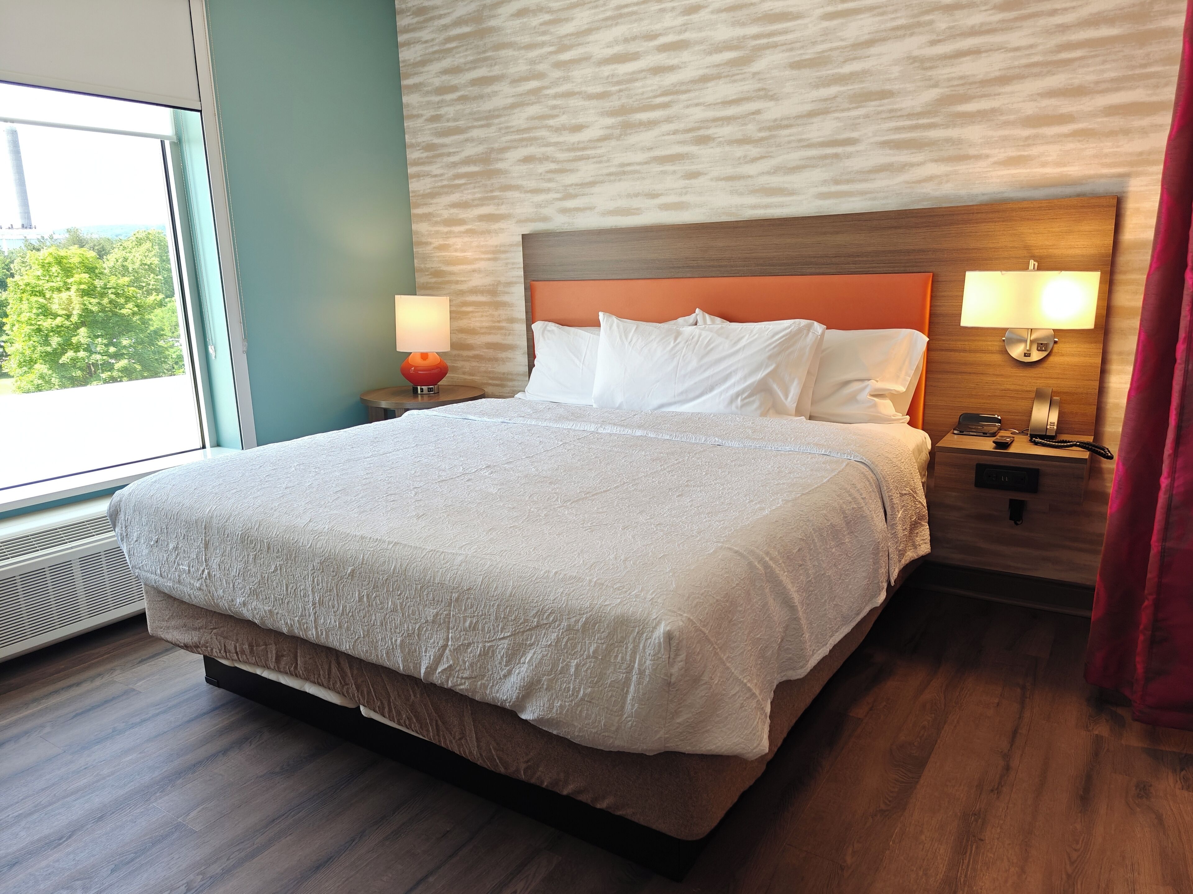 A hotel room with a large bed, two bedside lamps, a nightstand, and a window with a view of greenery.