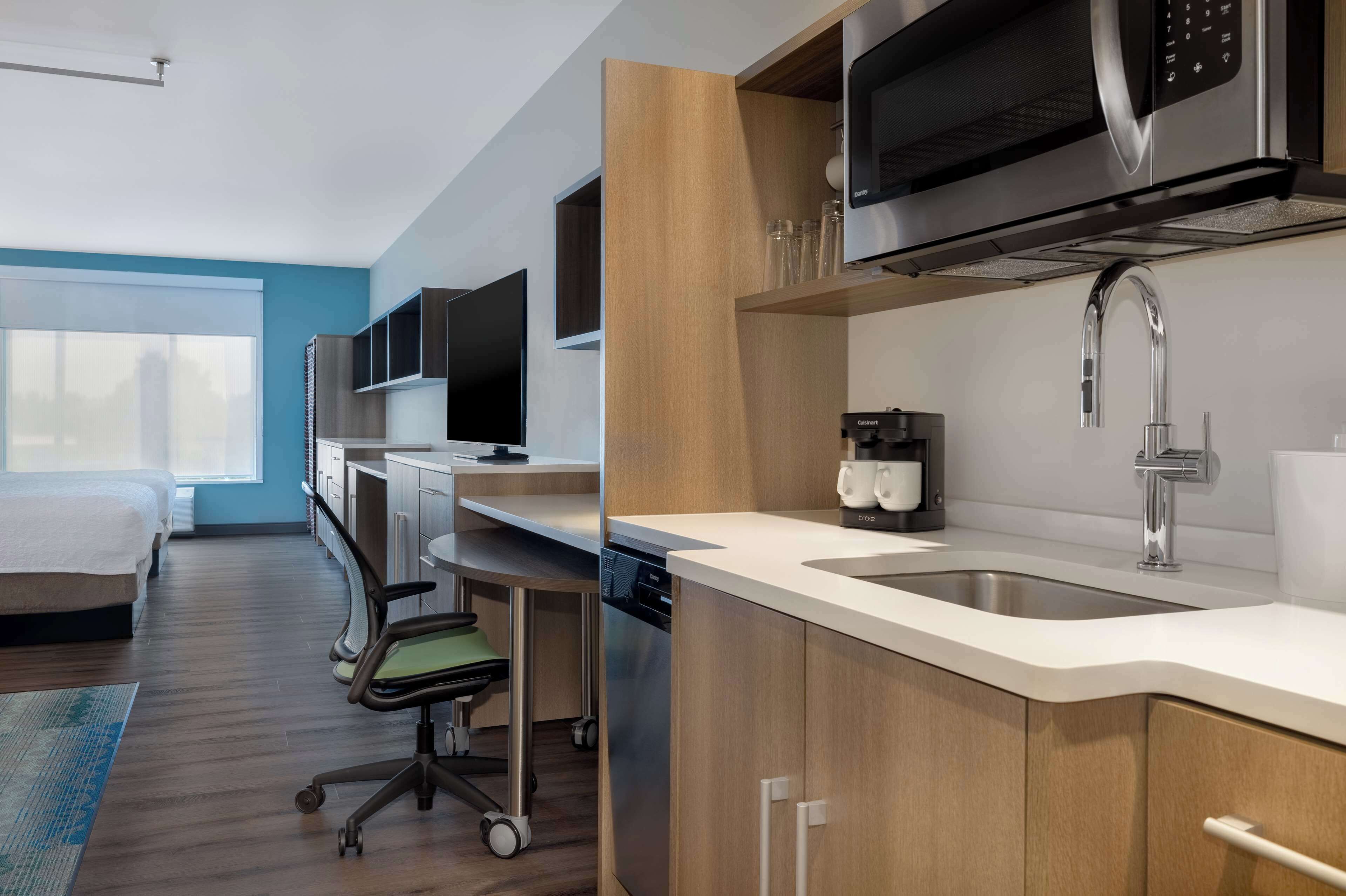 A modern kitchen with a microwave, sink, and coffee maker. There is a desk with a chair and a dishwasher.