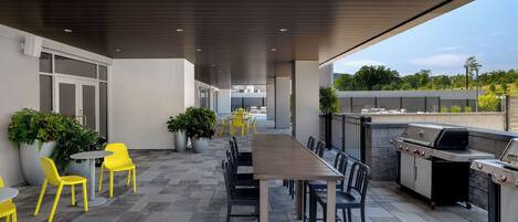 Un patio exterior con una mesa larga y sillas, una parrilla y plantas en macetas.