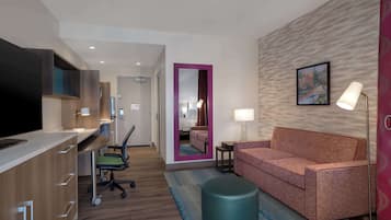 A hotel room with a kitchenette, a sofa, a desk with a chair, a mirror, a lamp, and a painting on the wall.