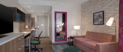 A hotel room with a kitchenette, a sofa, a desk with a chair, a mirror, a lamp, and a painting on the wall.