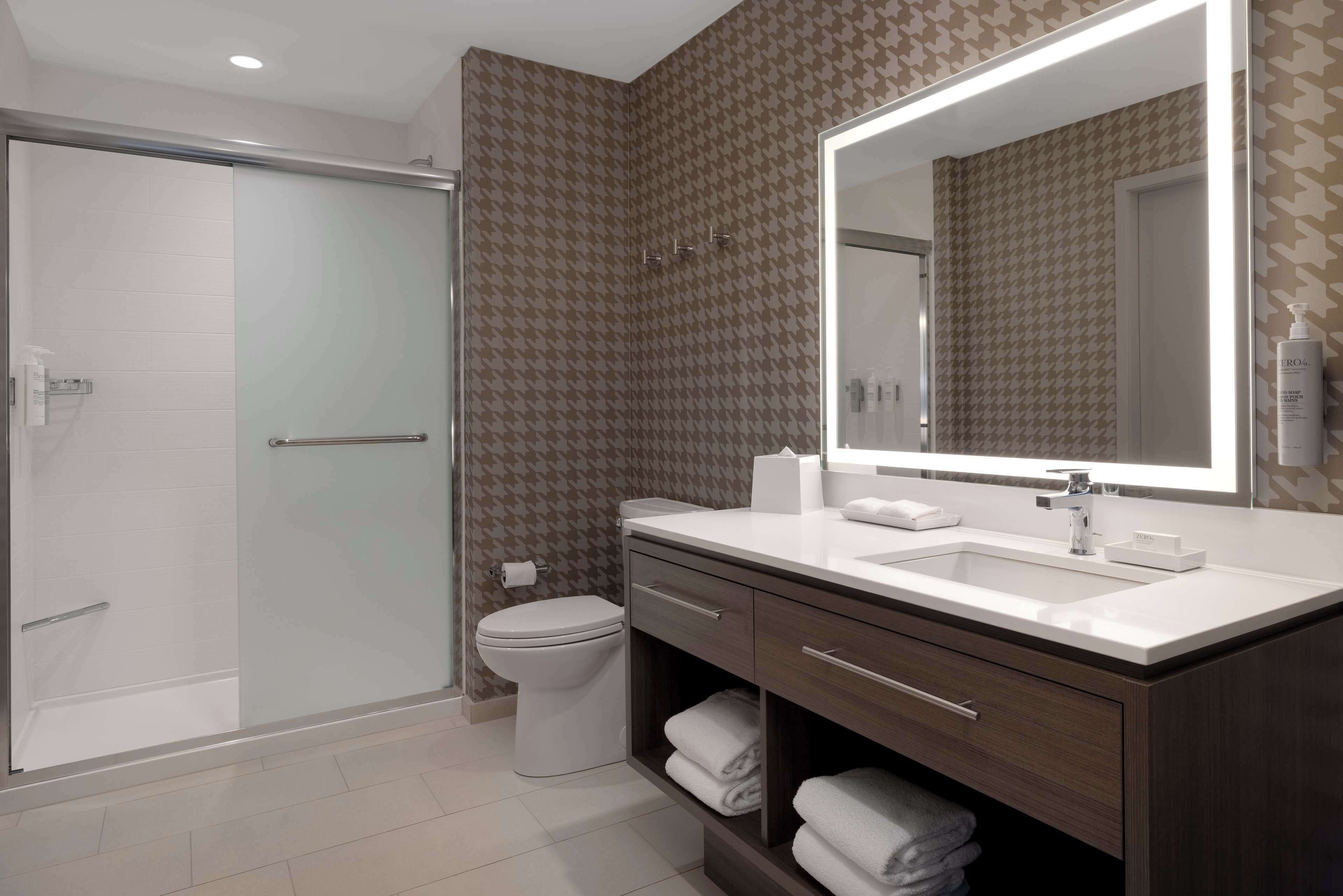 Une salle de bain moderne dotée d’une douche à parois de verre, de toilettes blanches, d’un grand miroir et d’un meuble-lavabo avec un espace pour ranger les serviettes.