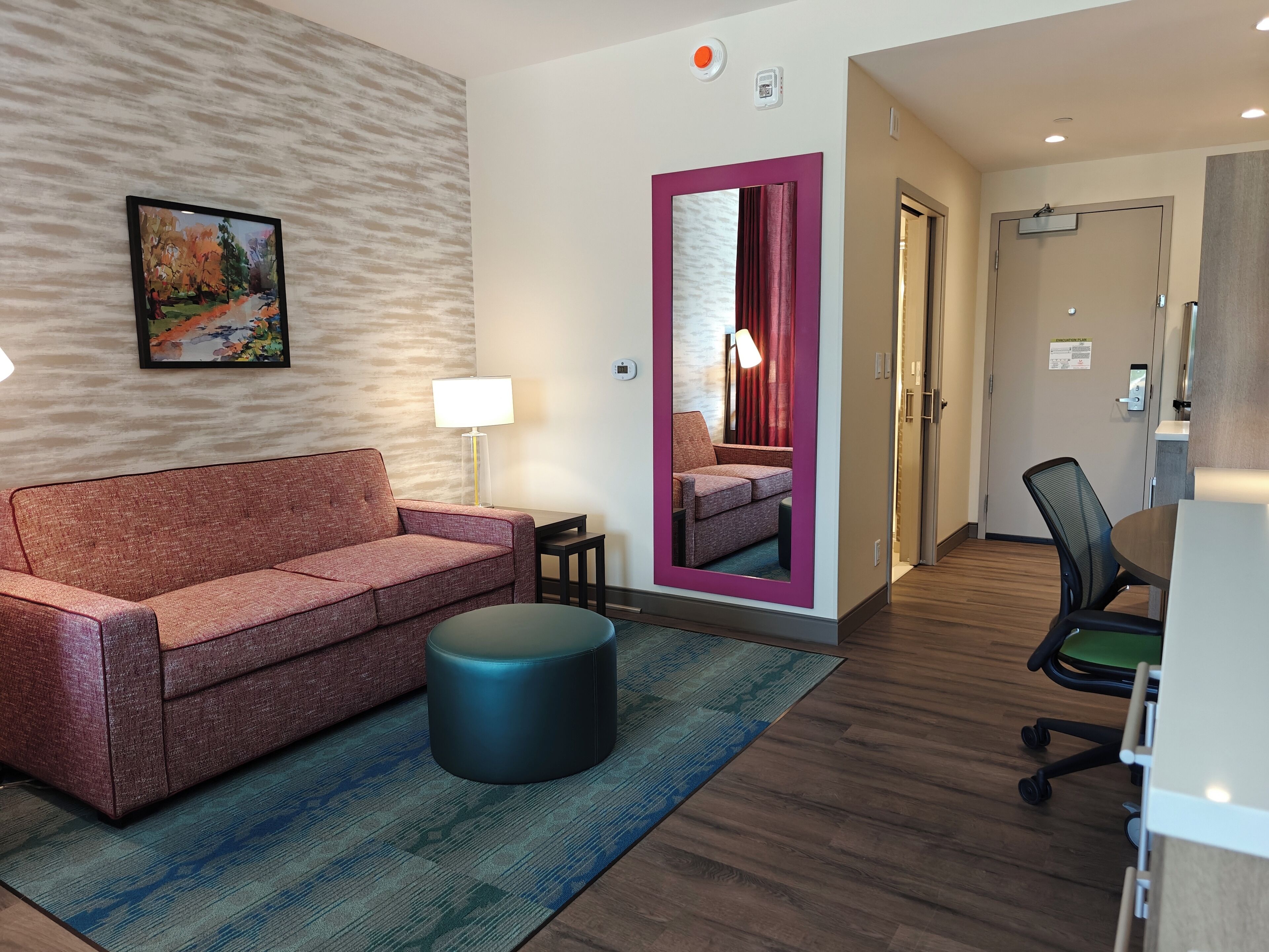 Una habitación de hotel con un sofá rojo, un taburete redondo pequeño, suelo de madera, un escritorio con silla, un espejo y un cuadro en la pared.