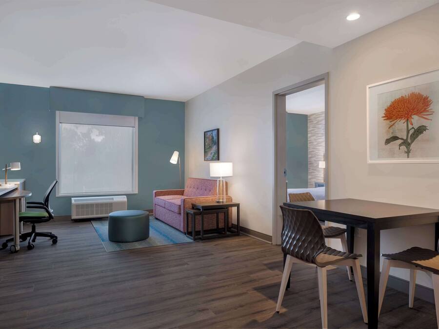 A modern hotel room with a desk, chair, sofa, and a dining table with chairs.