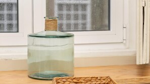 Une bouteille en verre transparent avec un bouchon tressé, posée sur une surface en bois, un panier tressé rempli de noix, et une fenêtre avec un motif quadrillé en arrière-plan.