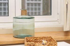 Eine klare Glasflasche mit Korkverschluss auf einer Holztischplatte, ein geflochtener Korb mit Nüssen und ein Fenster mit Gittermuster im Hintergrund.