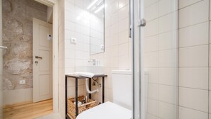 Une salle de bain avec des toilettes blanches, un lavabo posé sur un support en métal noir, un mur en pierre et un sol carrelé.