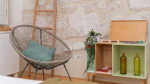 Une chaise en osier avec un coussin turquoise, une échelle en bois appuyée contre un mur de pierre, et une étagère verte sur laquelle se trouvent deux bouteilles et une boîte en bois.