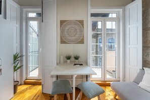 Un espace de vie compact comprenant une table blanche, deux chaises, une décoration murale et une vue sur la rue grâce à de grandes fenêtres.