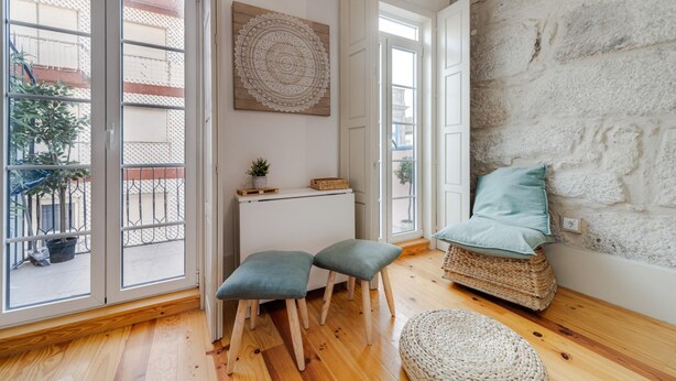 A room with wooden flooring, a stone wall, a wicker chair with a cushion, a small white cabinet, and two matching stools. - Bonjardim City Flats - Relaxing Terrace (Porto)