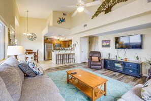 A spacious living room with a sofa, coffee table, and a TV. A kitchen is visible in the background with wooden cabinets and a dining area. - Mesquite Vacation Rental w/ Pool Access! (Mesquite)