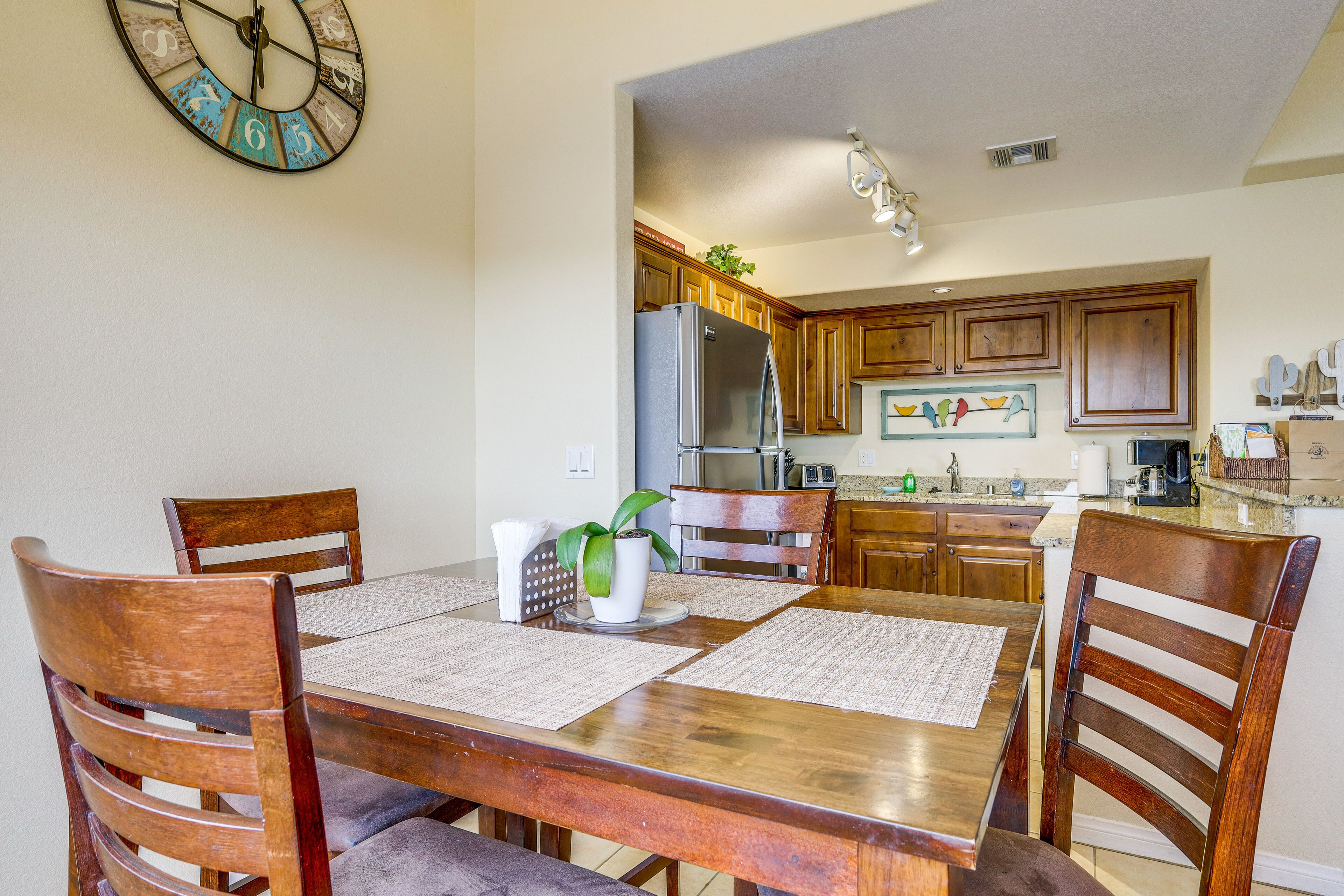 A dining area with a wooden table and chairs, a clock on the wall, and a kitchen in the background.