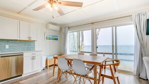 A spacious kitchen with a wooden dining table and chairs, a ceiling fan, and a view of the ocean through large windows. - Beachy Palms Cottage on Lake Huron w/ Hot Tub! (Palms)
