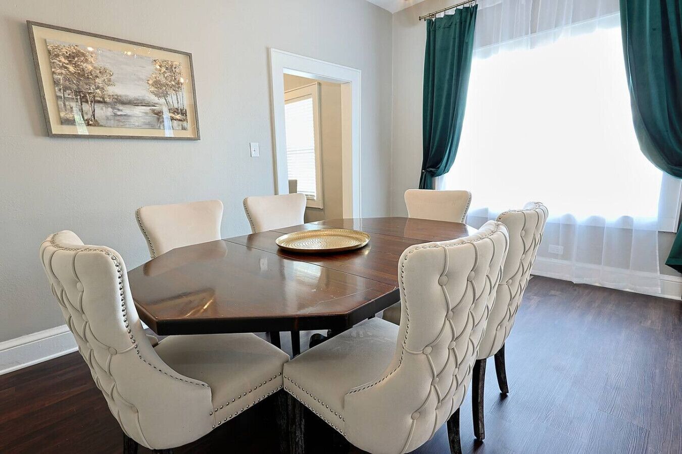 Une salle à manger avec une table ronde, six chaises rembourrées, un tableau encadré au mur et une fenêtre avec des rideaux.
