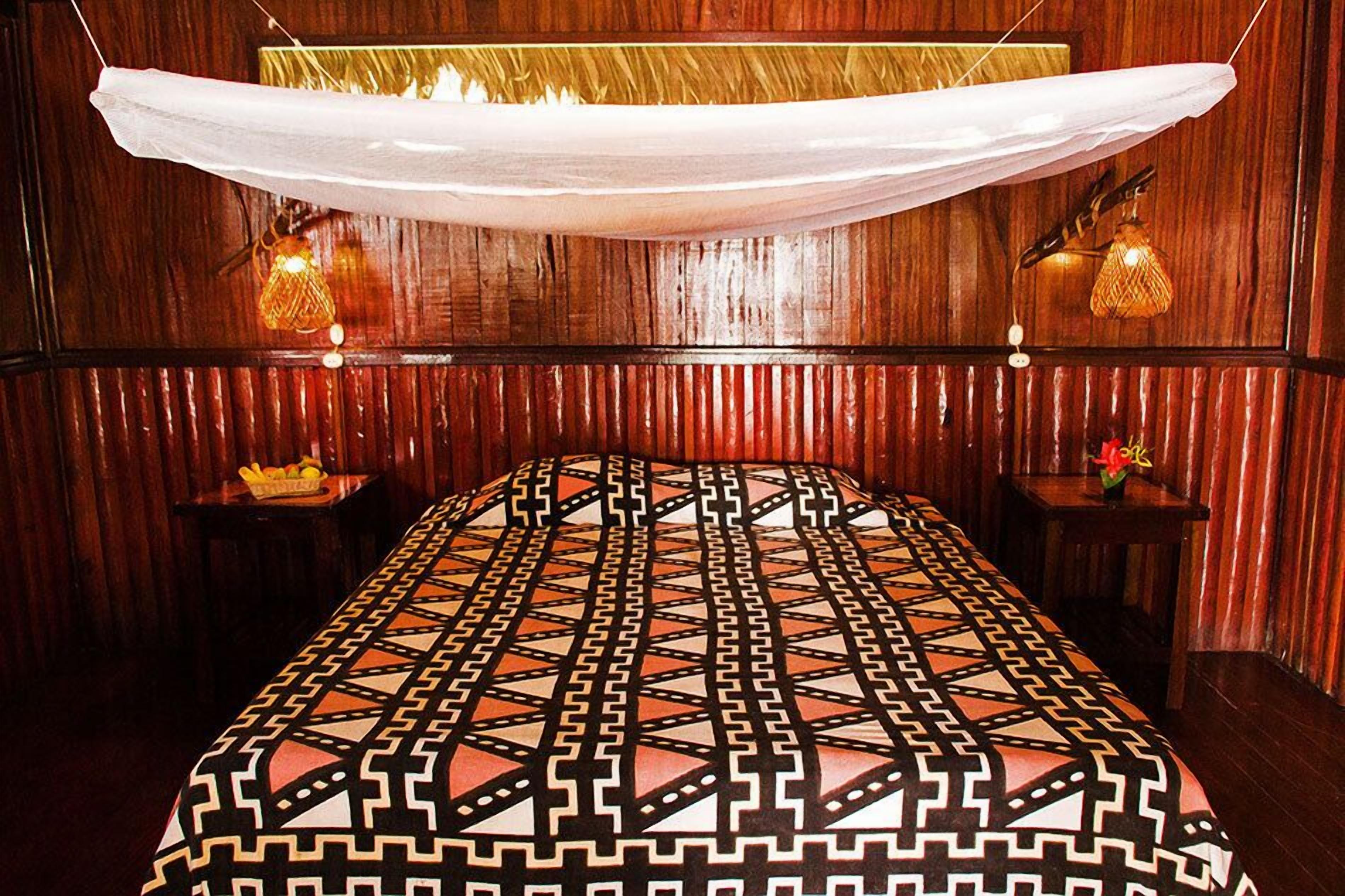 A room with a patterned bedspread, wooden panel walls, and two bedside tables with lamps.